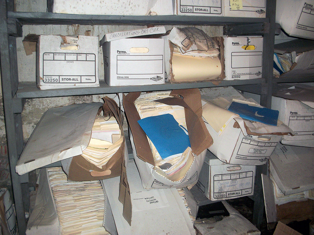 Water and mold damaged paper documents in cardboard bins on shelves in the basement